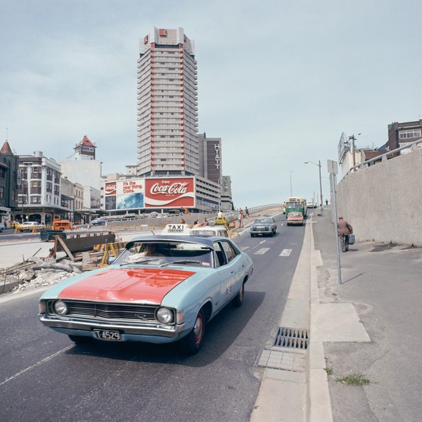A photo from the 1970s featuring an old blue and red taxi driving down a road. In the background is the iconic Coca Cola sign in Kings Cross Sydney